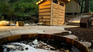 A wooden cabin with a small shed, surrounded by a stone fire pit and a wooden deck.