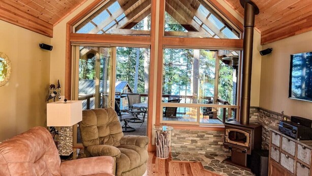 A cozy cabin interior with a large wooden ceiling, a stone fireplace, a comfortable leather couch, and a dining area visible through large windows.
