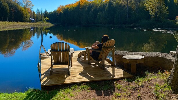 Um cais de madeira com duas cadeiras Adirondack e uma pequena mesa redonda, com vista para um lago tranquilo rodeado por árvores de cores outonais.