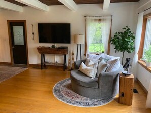 A living room with a grey sofa, a round rug, a wooden coffee table, a flat-screen TV, a potted plant, and a window with curtains.
