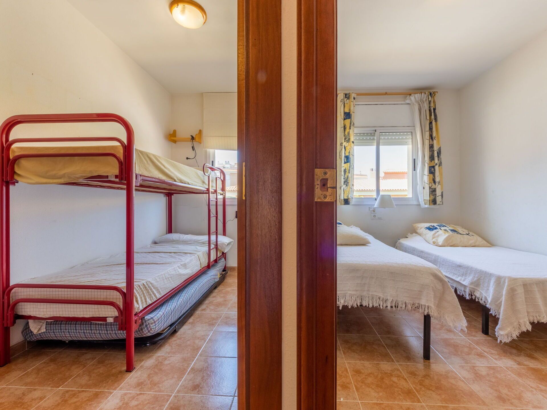 A dormitory room with bunk beds and two single beds, a window with curtains, and a tiled floor.