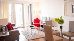 Ein modernes Wohnzimmer mit Sofa, Sessel, Glastisch und einem kleinen Beistelltisch mit Lampe.