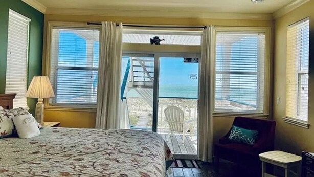 Ein Schlafzimmer mit Bett, Sessel, Lampe und Blick auf den Strand durch eine Glastür.