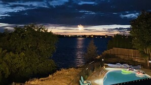 A swimming pool with lounge chairs, a wooden fence, and a view of a body of water with fireworks in the distance. - Spacious 4 bedroom lakefront house: with private dock and pool. (Moses Lake)