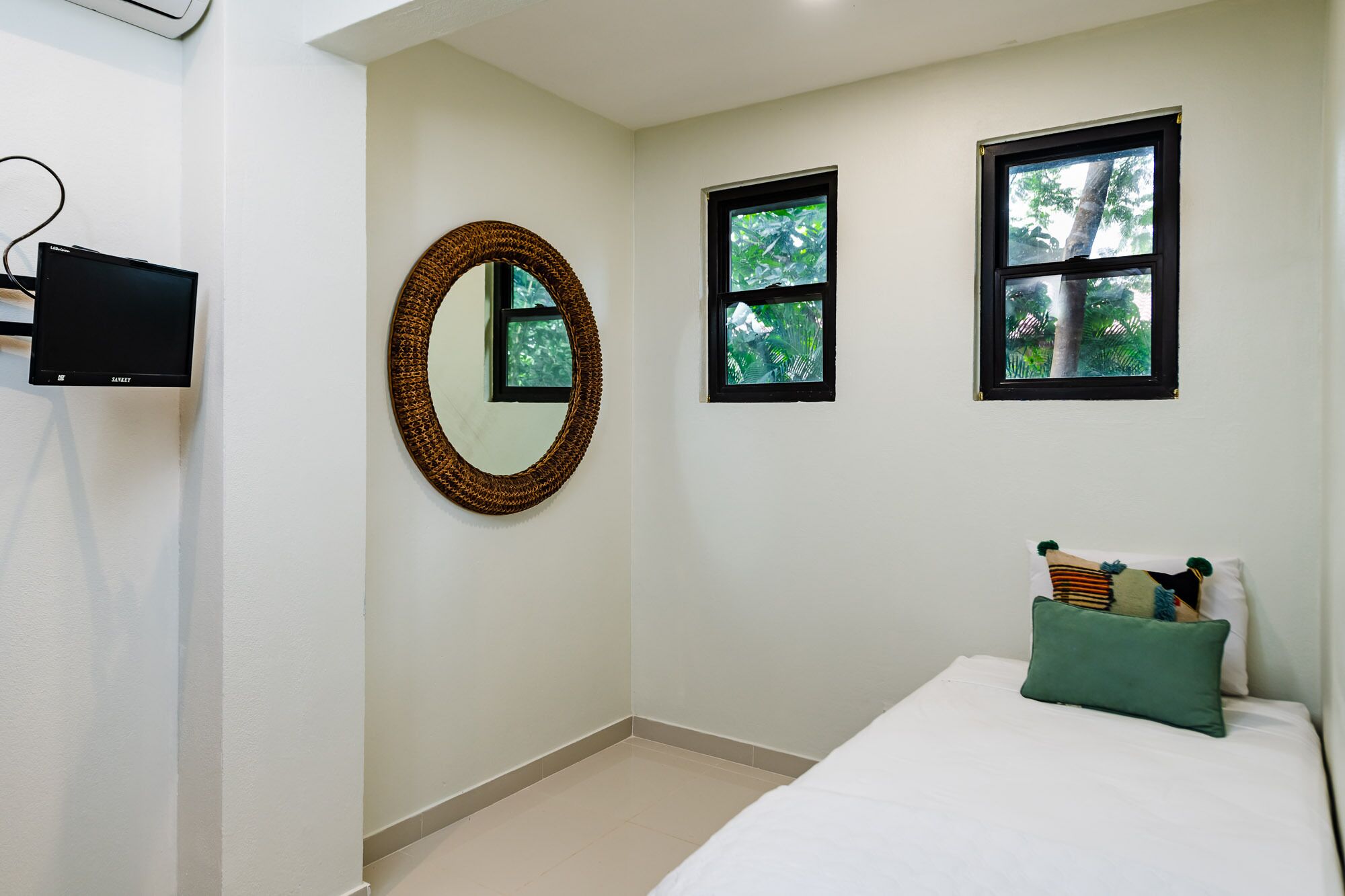 Une chambre à coucher comprenant un lit, un téléviseur fixé au mur, un miroir rond et deux fenêtres donnant sur des arbres.