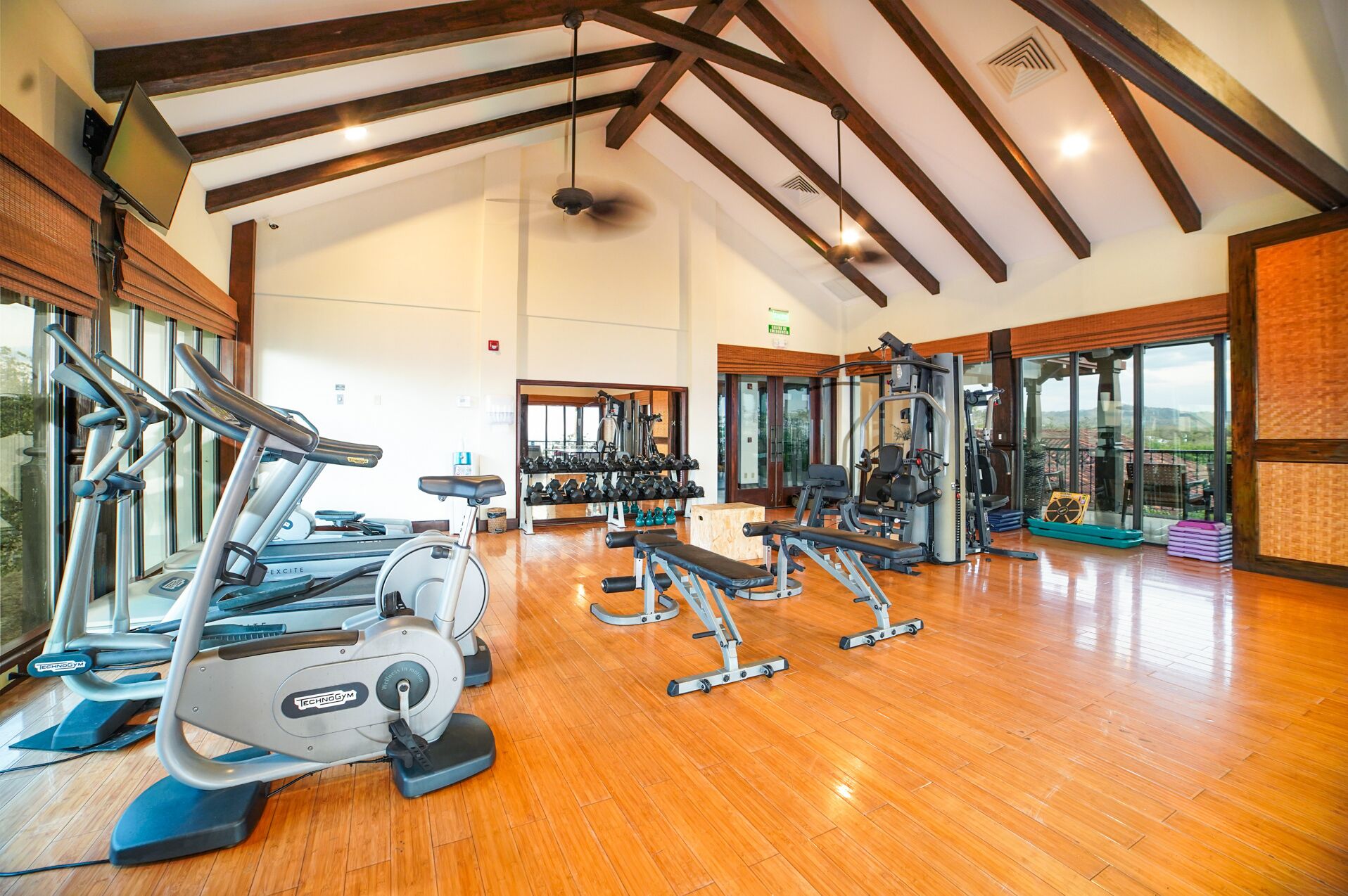 Une salle de sport spacieuse, dotée de divers appareils d’exercice, de grandes fenêtres et d’un plafond haut avec des poutres apparentes.