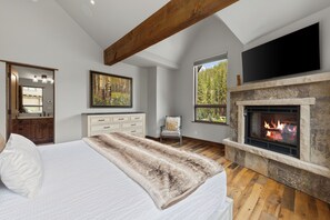 Un dormitorio con cama, chimenea, televisor, cómoda, espejo y ventana con vista al bosque.
