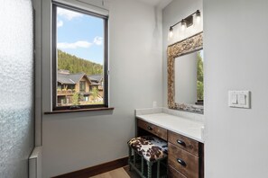 Un baño con ventana, espejo y un taburete de madera.