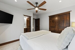 Une chambre à coucher comprenant un lit, un ventilateur de plafond, des armoires en bois, un téléviseur à écran plat et une porte donnant sur une autre pièce.