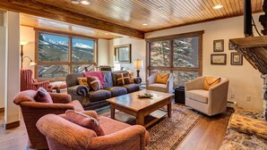 A cozy living room with a wooden ceiling, large windows with mountain views, a brown leather sofa, a patterned armchair, and a coffee table. - Amazing Views | Walk or Free Shuttle to Town (Breckenridge)