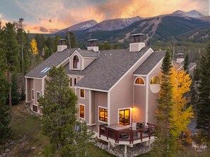 A modern two-story house with a deck, surrounded by trees and mountains.