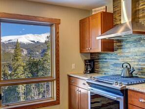 A kitchen with wooden cabinets, a stove, a coffee maker, and a view of snow-capped mountains through a window.