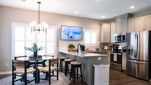 A modern kitchen with a central island, stainless steel appliances, and a dining area with a round table and chairs. - The Jubilee (Northport)