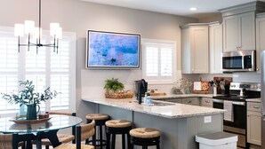 A modern kitchen with a central island, bar stools, a dining table, and a TV mounted on the wall. - The Jubilee (Northport)