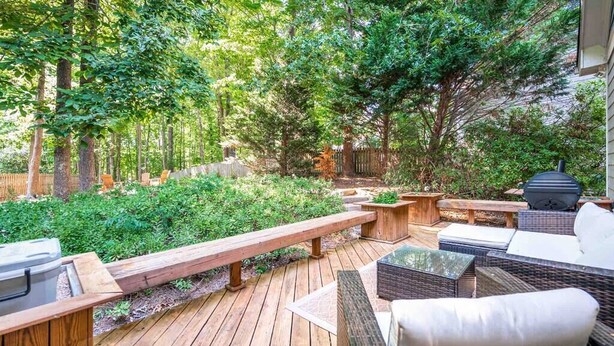 Une terrasse en bois, agréée de meubles d’extérieur, notamment un canapé et une table, et entourée d’une végétation luxuriante et d’arbres.