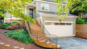 Une maison à deux étages avec un escalier en briques menant à la porte d’entrée, un garage et un jardin bien entretenu, agrémenté de paillis et d’arbustes.