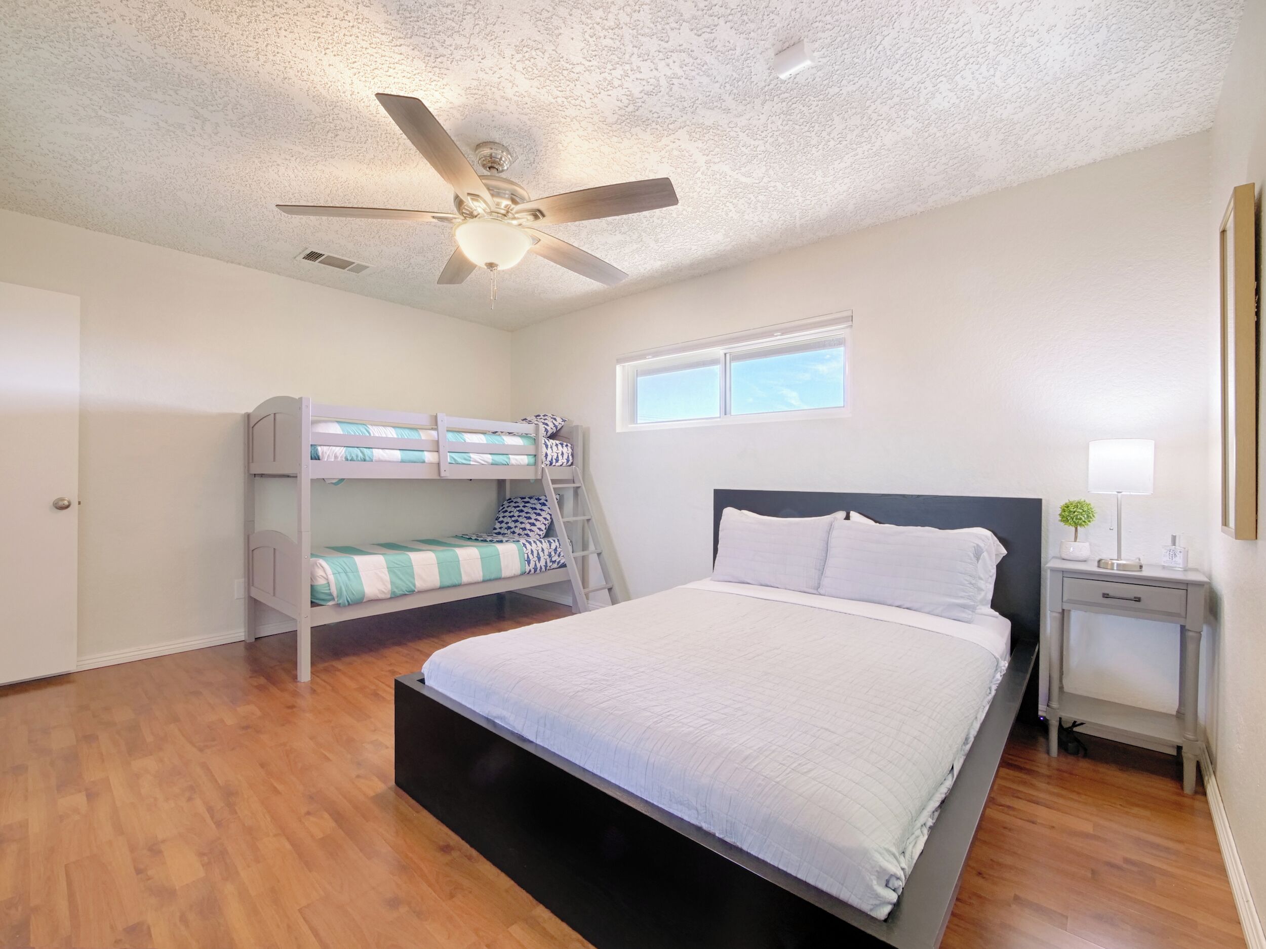 A bedroom with a bunk bed, a single bed, a ceiling fan, a nightstand with a lamp, and a window.
