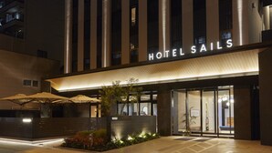 A modern hotel entrance with a sign reading "HOTEL SAILS," featuring a well-lit facade, outdoor seating with umbrellas, and a landscaped area with plants.