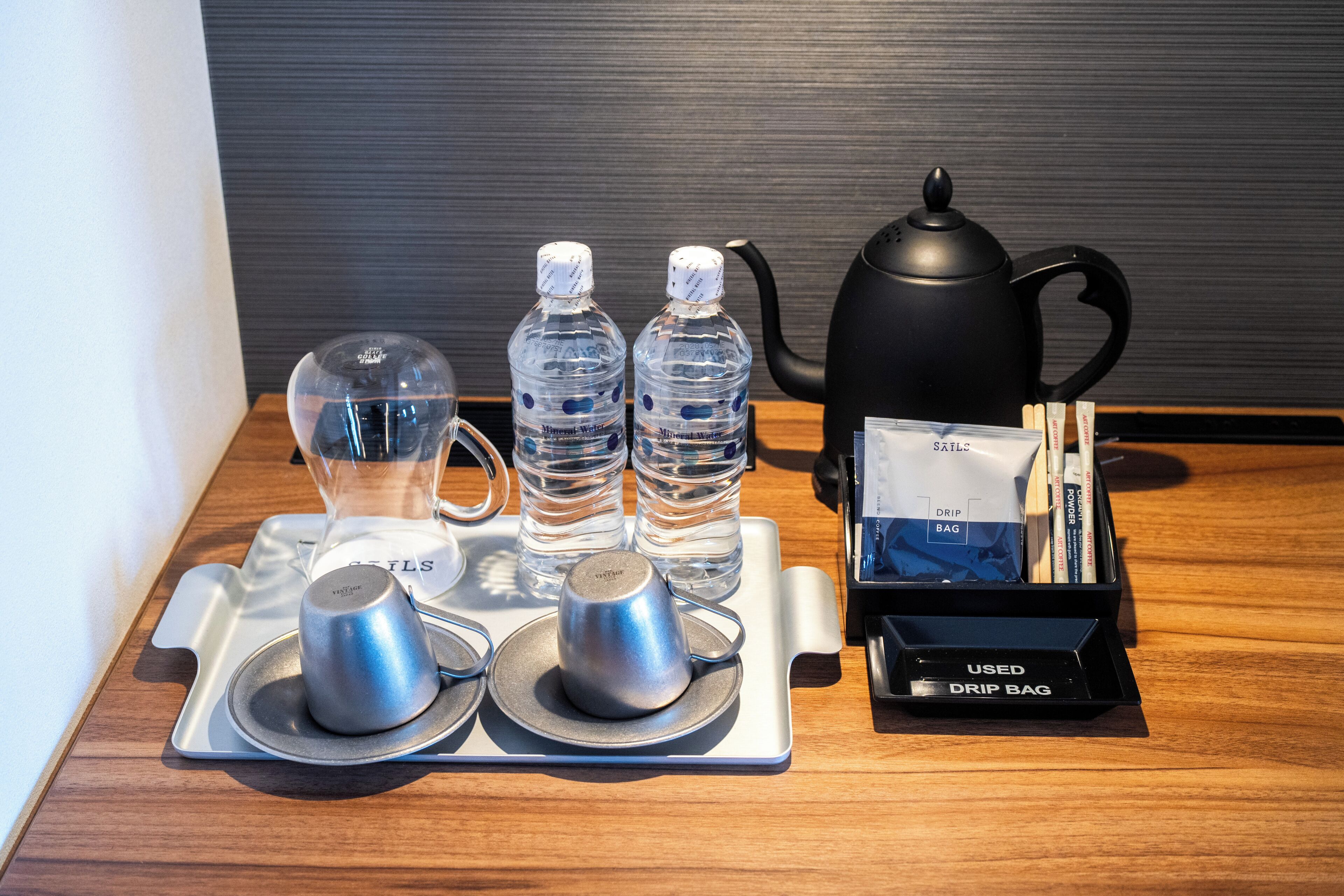 Un plateau contenant une théière, deux bouteilles d’eau, un pichet en verre et un sachet pour préparer le café, le tout posé sur une surface en bois.