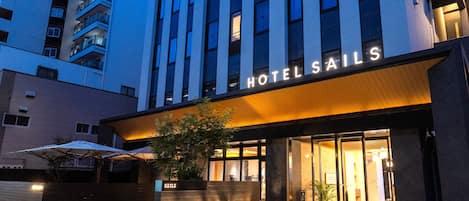 A hotel entrance with a sign reading 'HOTEL SAILS,' a glass door, and a small outdoor area with plants and an umbrella.