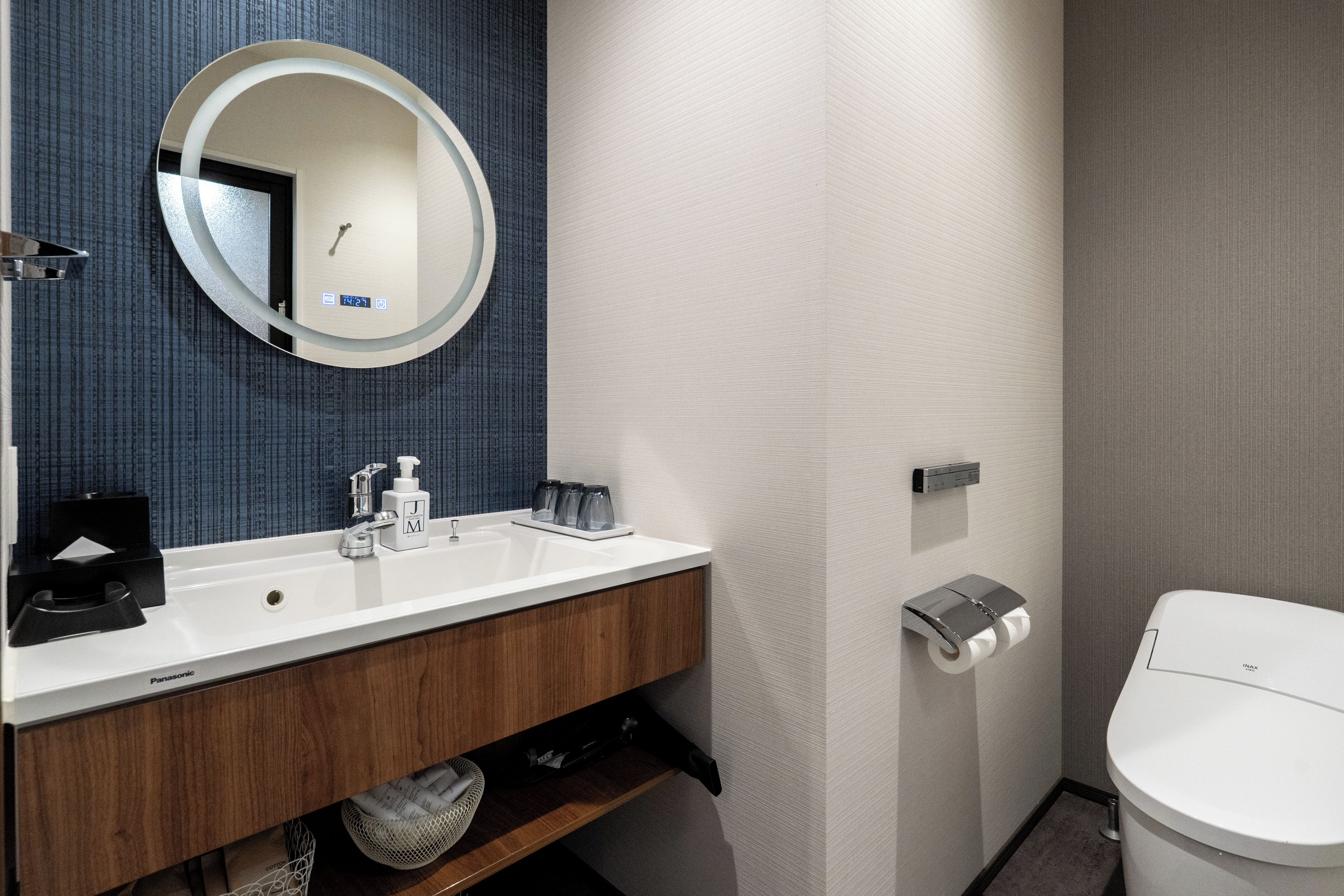 A modern bathroom with a round mirror, a sink with a soap dispenser, a toilet, and a paper towel dispenser.