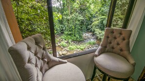 A room with a large window overlooking a lush garden. There is a tufted armchair and a cushioned chair near the window.