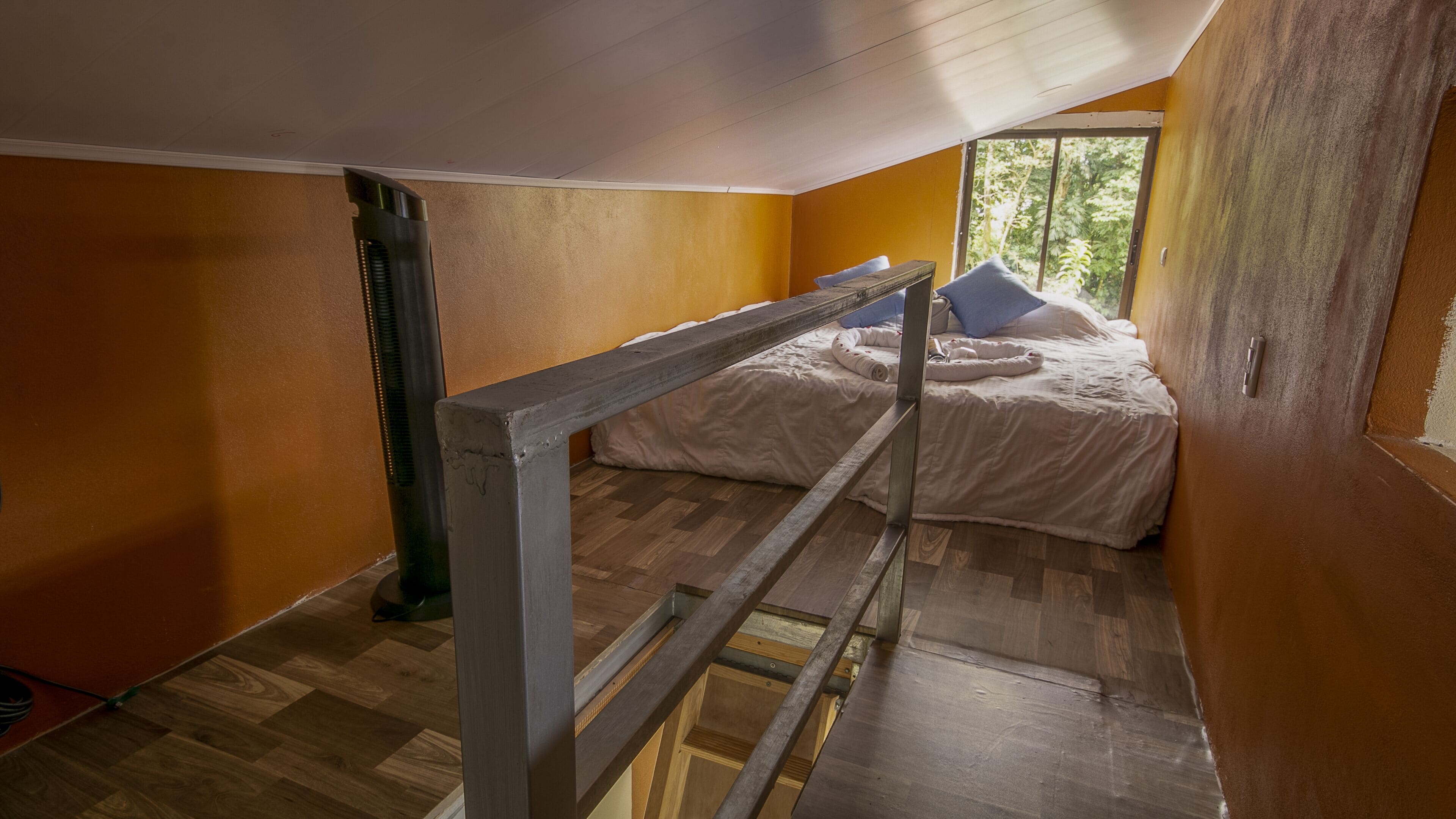 A bedroom with a wooden floor, a bed with white bedding, a window with a view of trees, and a wooden ladder.