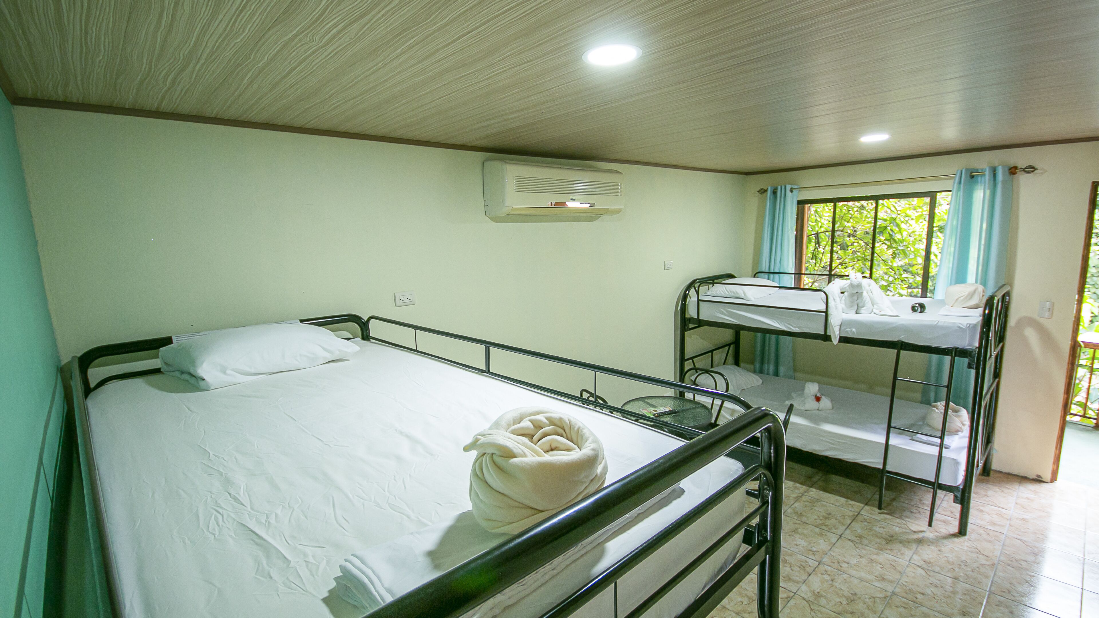 Ein Einzelzimmer mit einem Etagenbett im Hintergrund, einem Deckenventilator und einem Fenster mit Vorhängen.