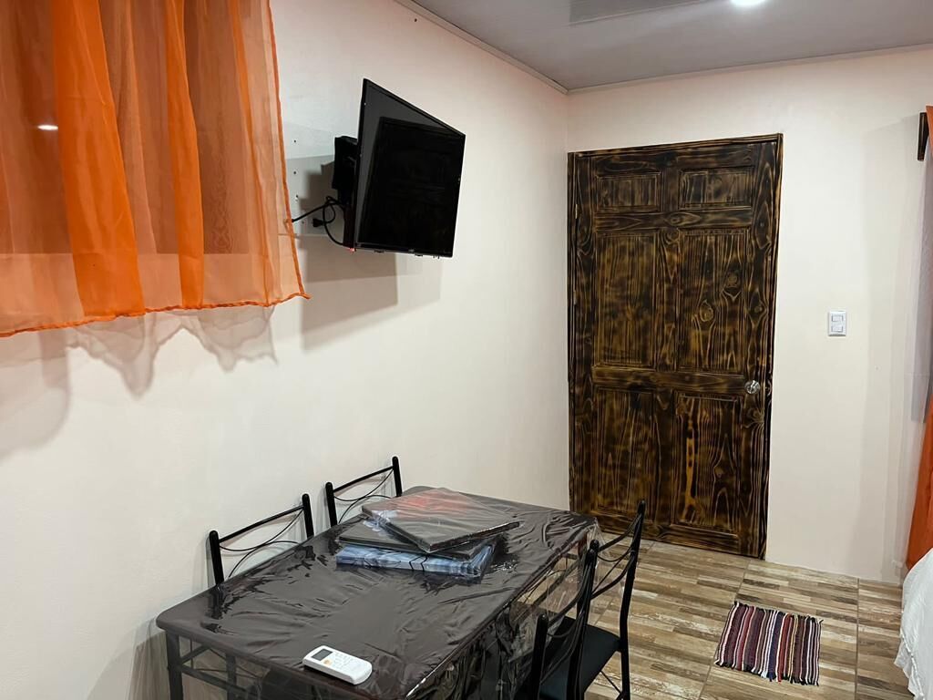 Una habitación con una mesa de comedor cubierta con plástico, un televisor montado en la pared y una puerta de madera.