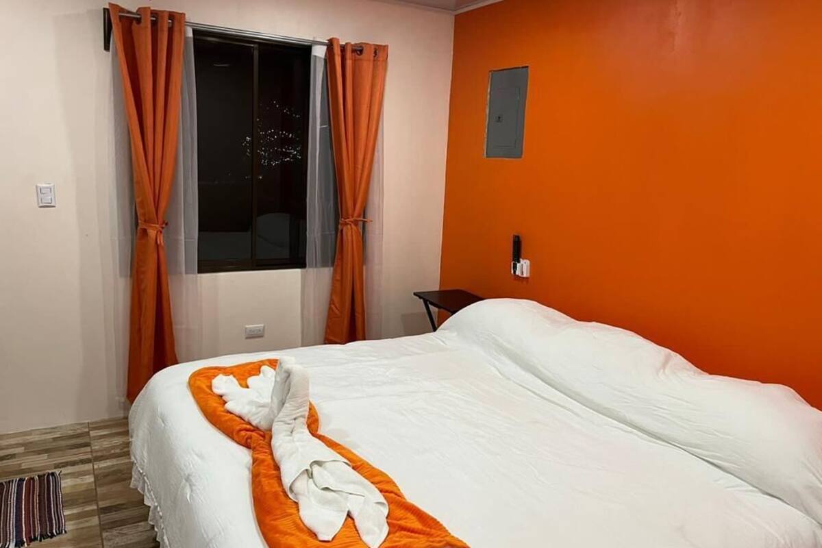 Una camera d'albergo con un letto, tende arancioni, una finestra e un interruttore luce a parete.