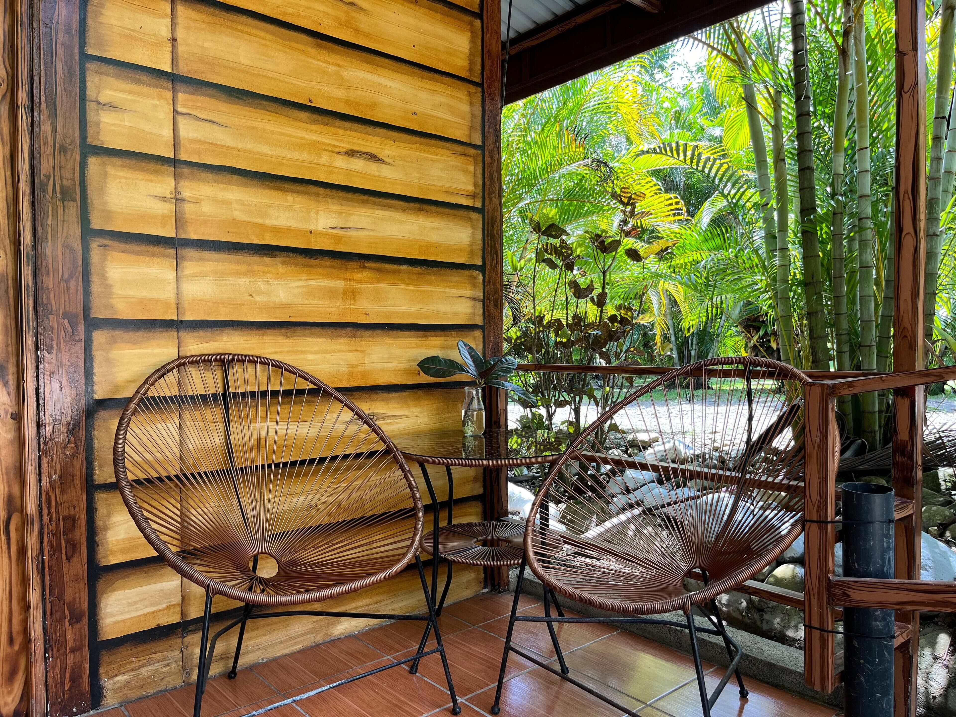 Un interior de madera con dos sillones de mimbre y una mesita, una pared de madera y vistas a un follaje exuberante.