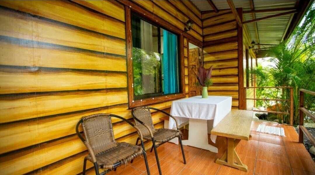 A wooden cabin with a table and two wicker chairs, a window with a curtain, and a view of greenery outside.