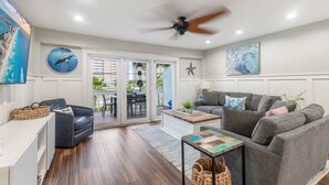 A spacious living area with a grey sofa, a coffee table, and a TV mounted on the wall. There is a ceiling fan, a large window, and a view of the outside. -  Beach. Walk everywhere. Peaceful. Location. (Navarre)