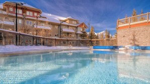 A clear pool surrounded by buildings with balconies, a stone wall, and snow-covered trees. - APEX Magnificent mountain view condo Pool &Hot Tub (Canmore)