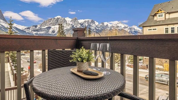 A balcony with a wicker table, a potted plant, and two wine glasses. - APEX Magnificent mountain view condo Pool &Hot Tub (Canmore)