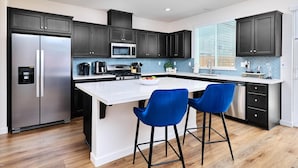 Een moderne keuken met donkere kastjes, roestvrijstalen apparatuur, een wit kookeiland en blauwe barstoelen.