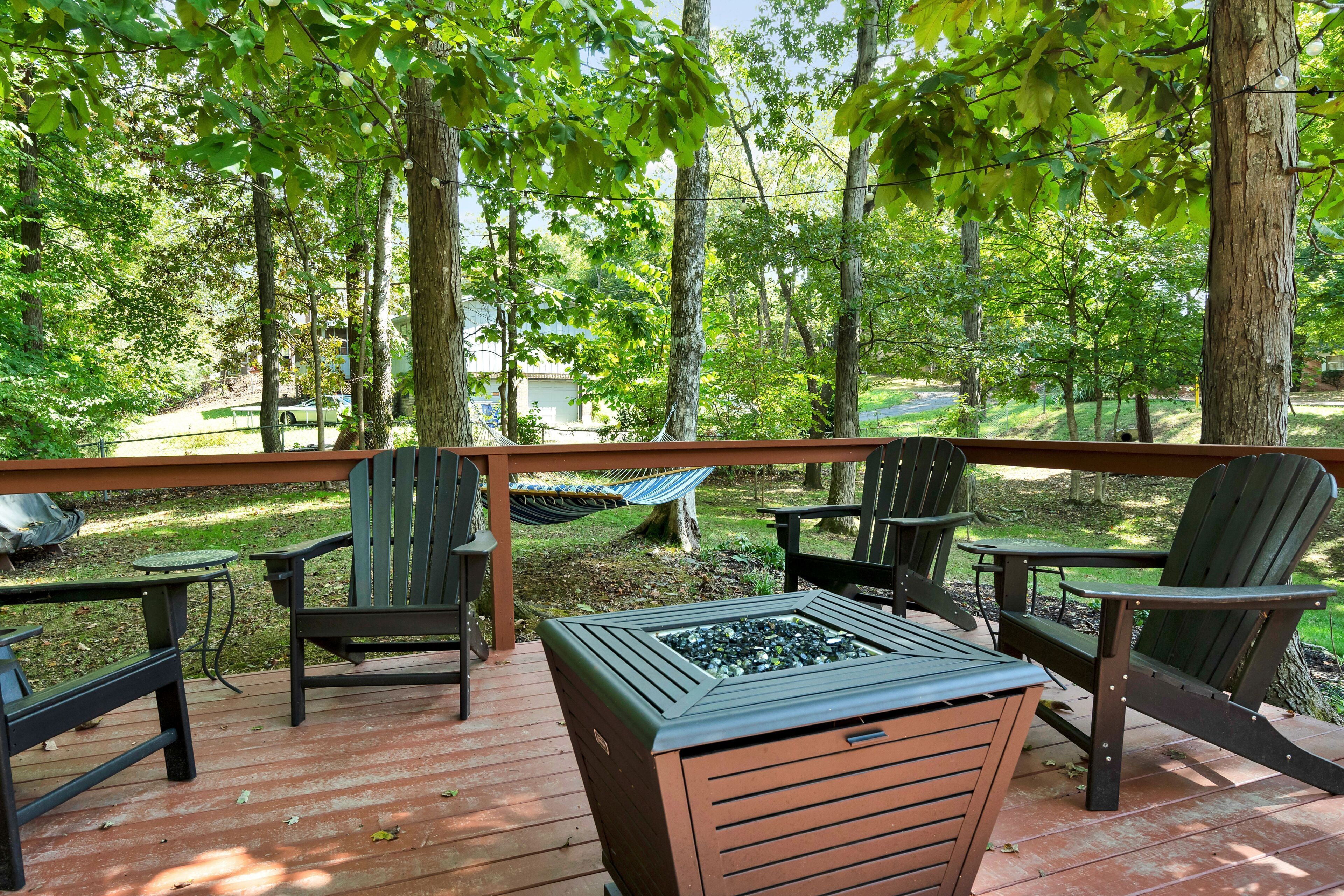 An outdoor deck with wooden flooring, surrounded by trees, featuring two Adirondack chairs and a small table with a glass top.