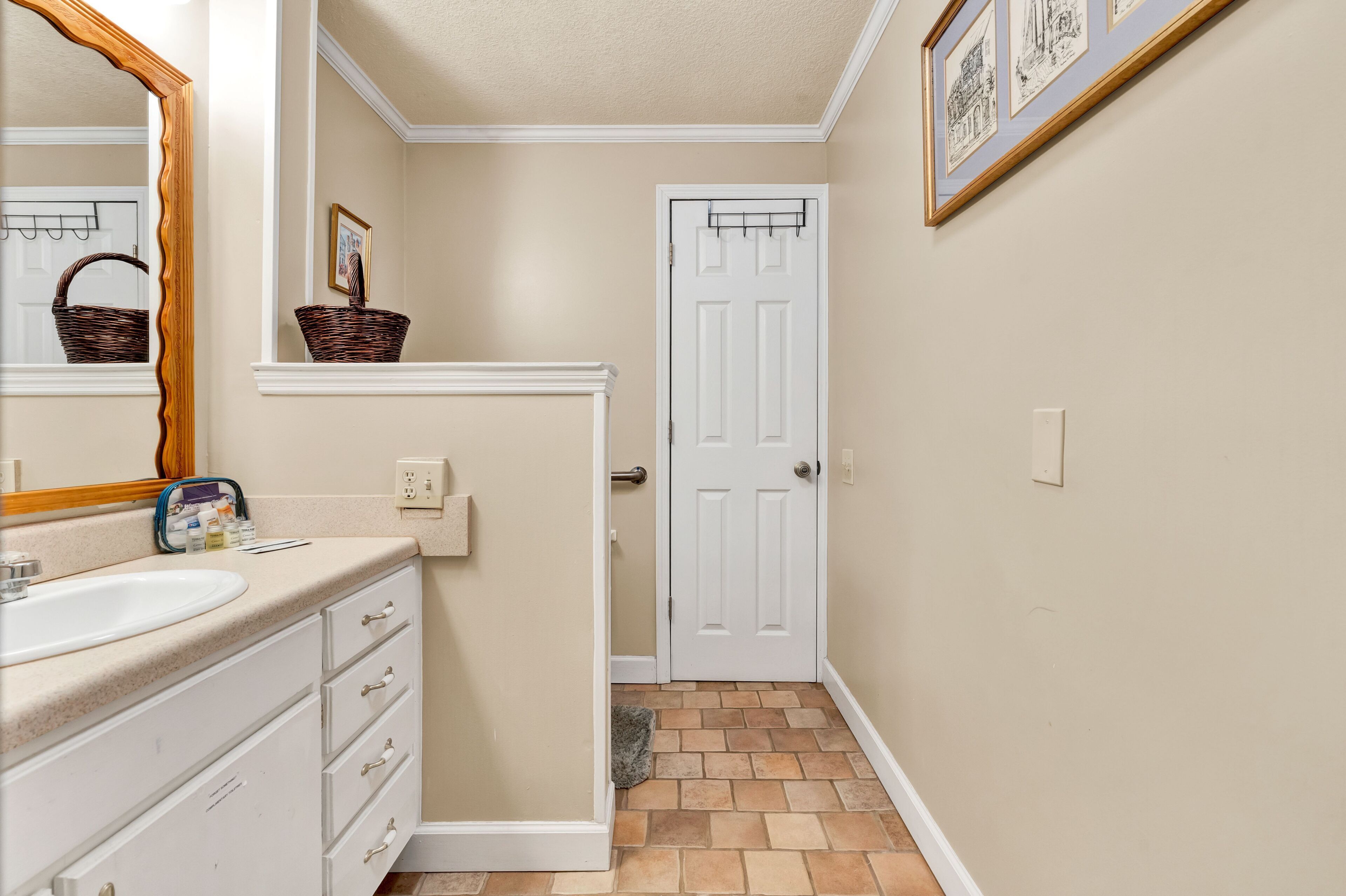 Une salle de bain avec un lavabo, un meuble-lavabo et un miroir blancs. On y trouve un panier sur le meuble-lavabo et une porte qui donne sur une autre pièce.
