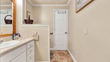 Une salle de bain avec un lavabo, un meuble-lavabo et un miroir blancs. On y trouve un panier sur le meuble-lavabo et une porte qui donne sur une autre pièce.
