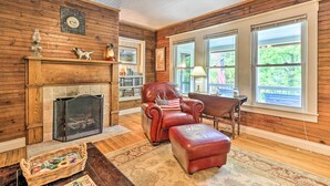 A cozy living room with a fireplace, leather armchair, ottoman, and large windows offering a view of the outdoors. - Fish, Float & Relax Fireside: Home Near New River! (Crumpler)