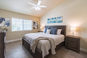 Ein Schlafzimmer mit Bett, Nachttisch, Deckenventilator, Fenster mit Jalousien und Bildern an der Wand.