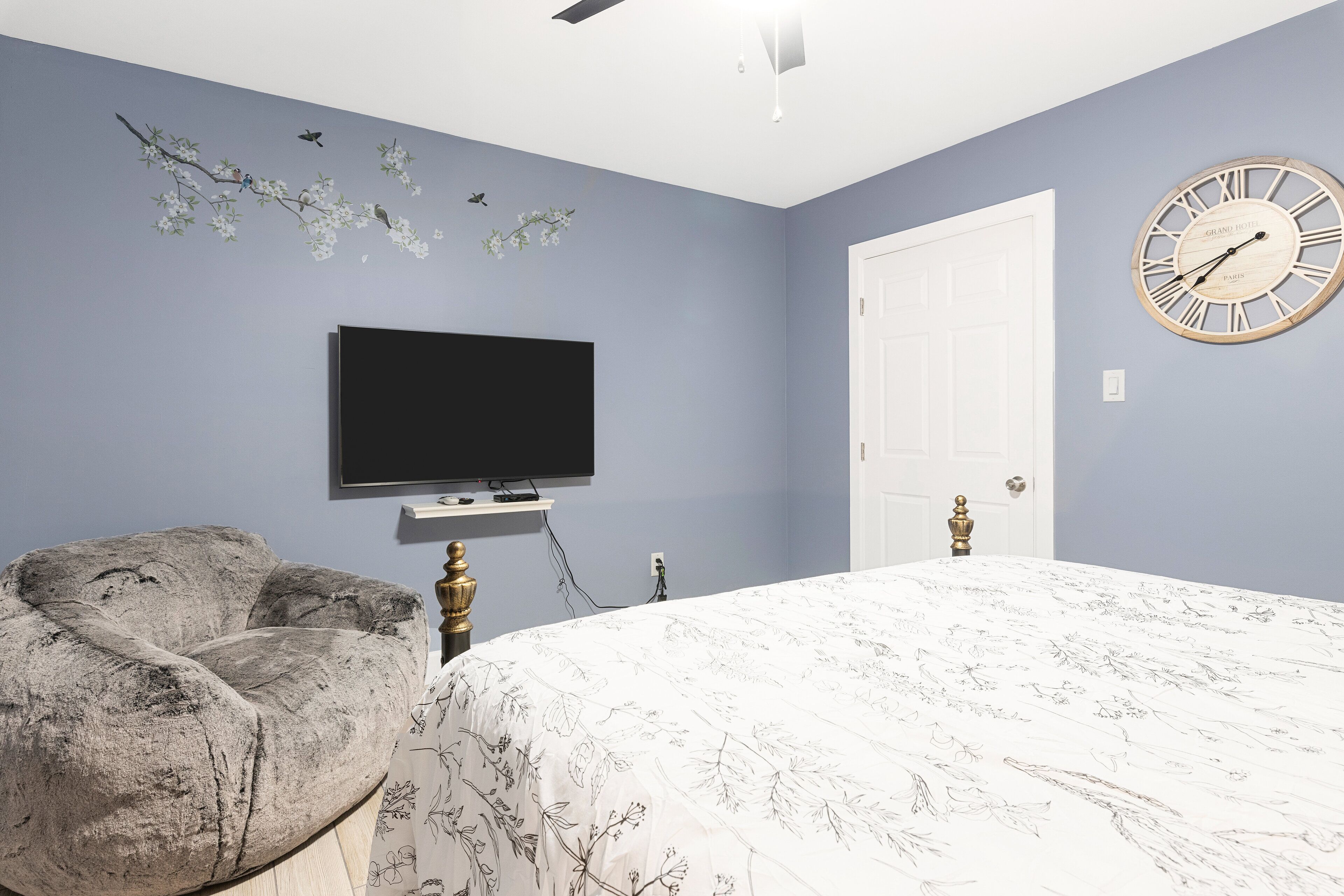 A bedroom with a bed, a flat-screen TV, a decorative wall with a branch and birds, a clock, and a door.