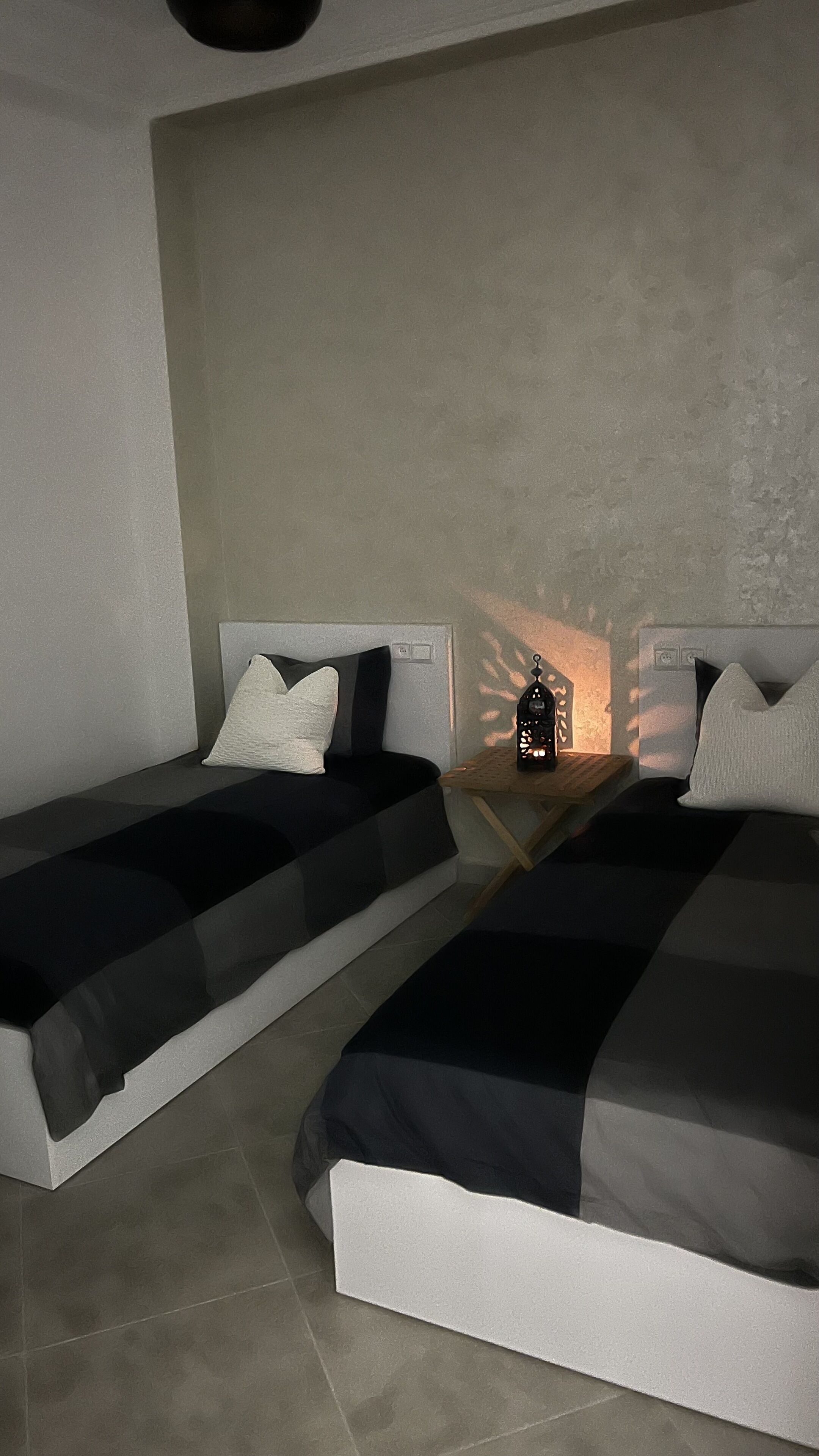 Una habitación pequeña y moderna con dos camas individuales, una mesita de noche con un farol y una pared lisa.