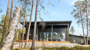 Moderni mökki, jossa on suuret ikkunat, ja se on ympäröity puilla. Lisäksi siinä on puinen terassi.
