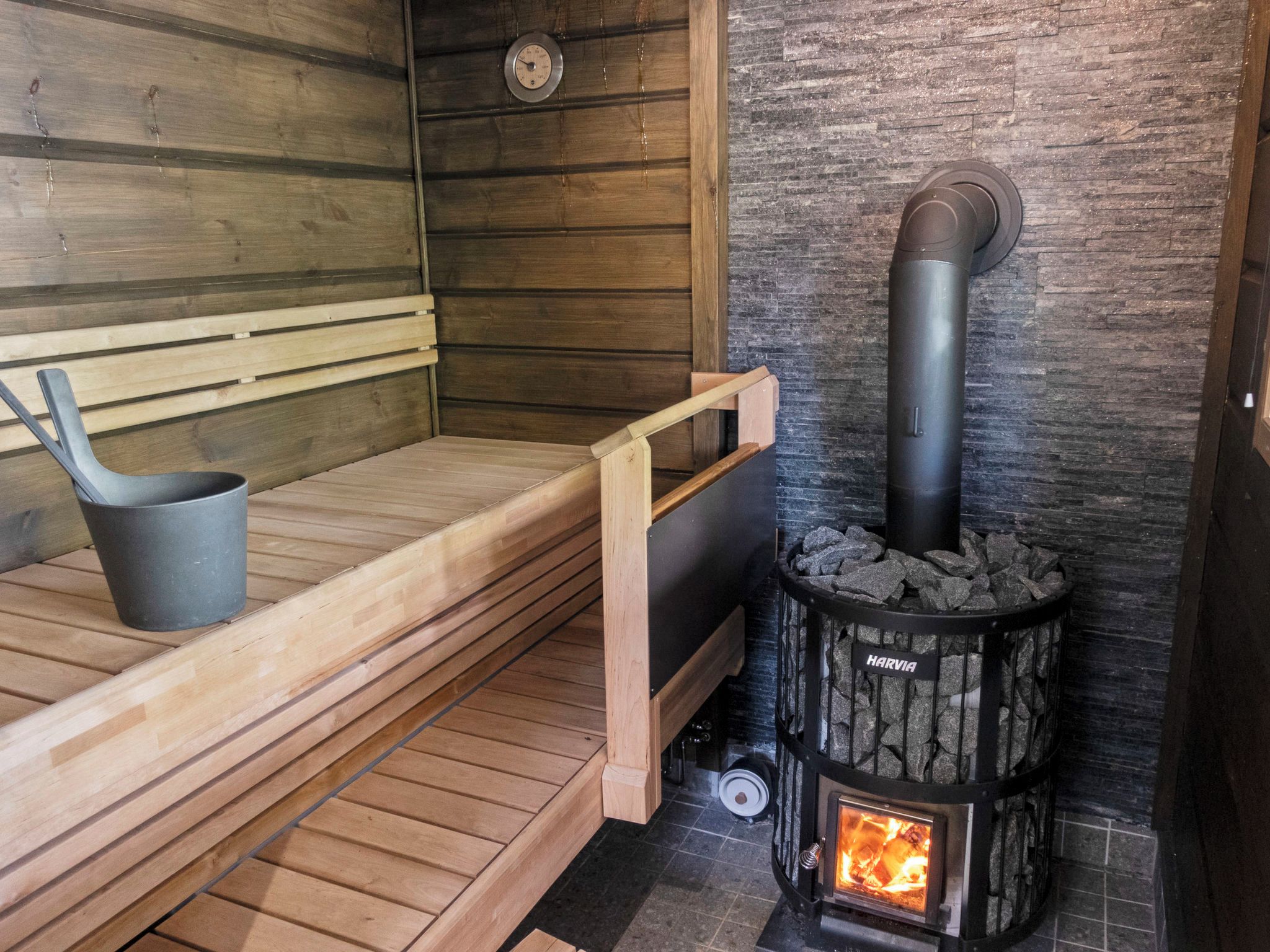 A sauna with a wooden bench, a black heater, and a stone wall.