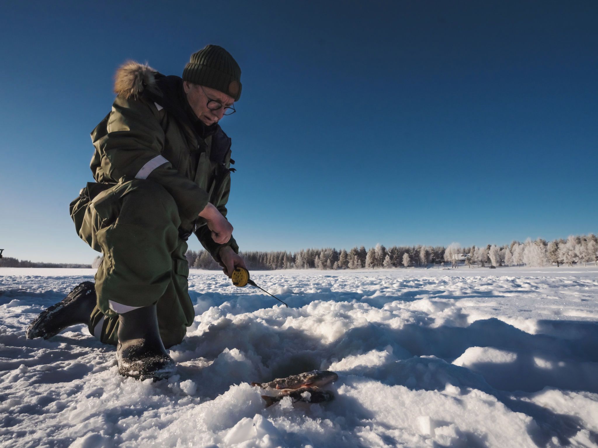 A person crouching on a snowy surface, holding a fishing rod.