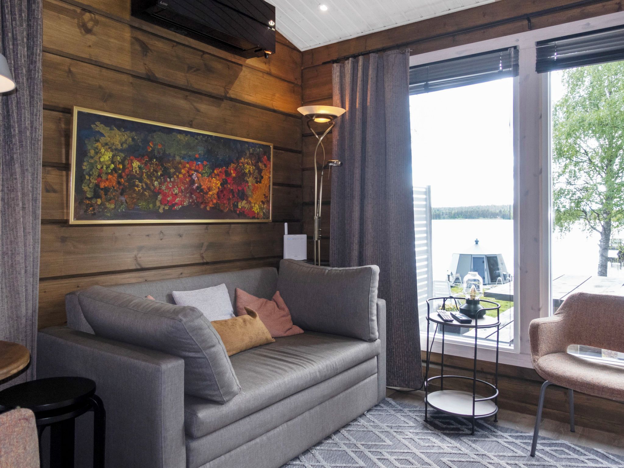A modern living room with a grey sofa, a painting, a floor lamp, and a view of a body of water through large windows.
