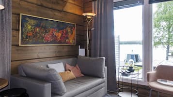Ein modernes Wohnzimmer mit einem grauen Sofa, einem Gemälde, einer Stehlampe und Blick auf ein Gewässer durch große Fenster.