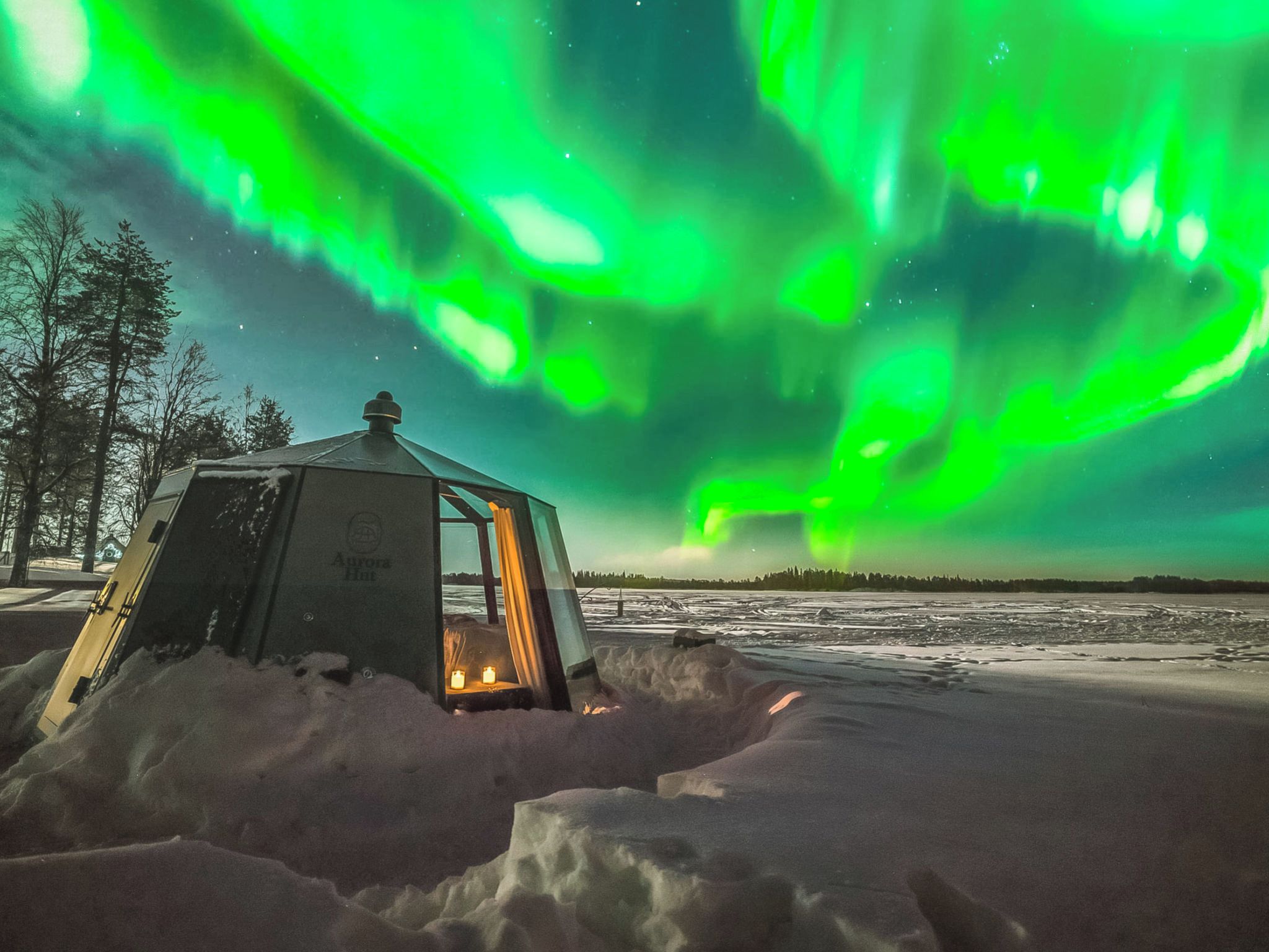 Un paysage enneigé avec une petite structure illuminée, qui pourrait être une cabane, et des aurores boréales en arrière-plan.