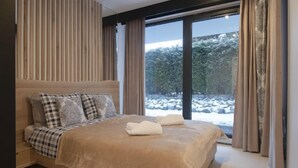 Ein modernes Schlafzimmer mit einem großen Bett, einem Kopfteil mit vertikalen Lamellen und Blick auf eine verschneite Landschaft durch große Fenster.